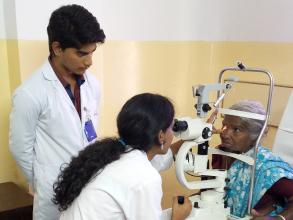 Auf dem Bild befinden sich ein junger Arzt und eine junge Ärztin, die eine Patientin untersuchen. Die Patientin sitzt auf einem Stuhl vor einem Sehtestgerät. Die Ärztin ist dabei, die Patientin auszumessen, während der andere Arzt ihr bei der Arbeit über die Schulter sieht.