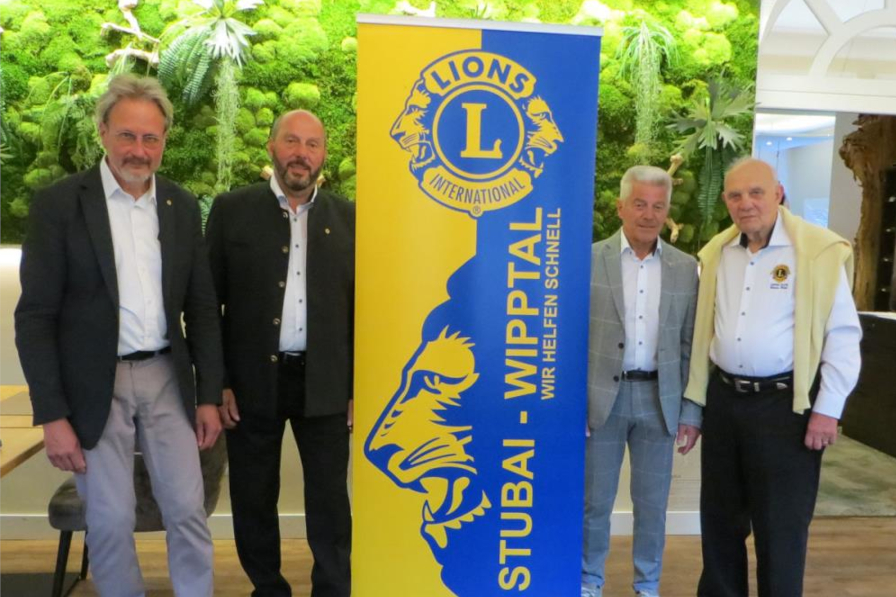 Das Bild zeigt 4 ältere Herren aus den beiden Lions Clubs. Sie stehen um einen Banner der Lions International der in den typischen Farben Gelb und Blau gehalten ist. Auf dem Banner steht "Stubai - Wipptal" der Hintergrund besteht aus einer grünen Pflanzwand.