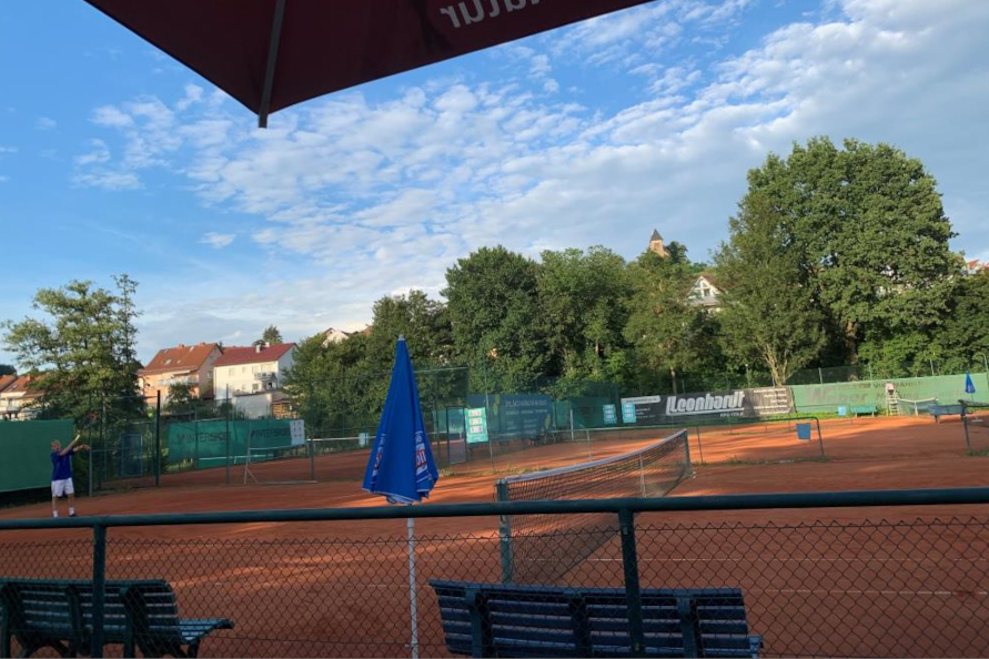 Ausblick vom Spielfeldrand auf das Tennisfeld. Es ist sonnig, mit blauem Himmel. Links weiter hinten sieht man einen Mann mit dunkelblauem Sportshirt und weißen Tennishosen, der gerade einen Aufschlag macht.