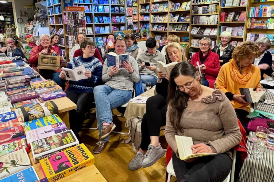 Auf diesem Bild sieht man die Gruppe Frauen aus einer anderen Perspektive. Die Gruppe umfasst ca. 15 Teilnehmerinnen und man sieht sie wieder lesen, sich miteinander unterhalten oder auf die Lesung vorbereiten. Den Laden sieht man nun aus einer anderen Perspektive und kann erkennen, dass im Vordergrund ein Tisch mit angebotenen Büchern steht und der Laden aus weiteren, deckenhohen Bücherregalen besteht die alle sehr gut gefüllt sind.