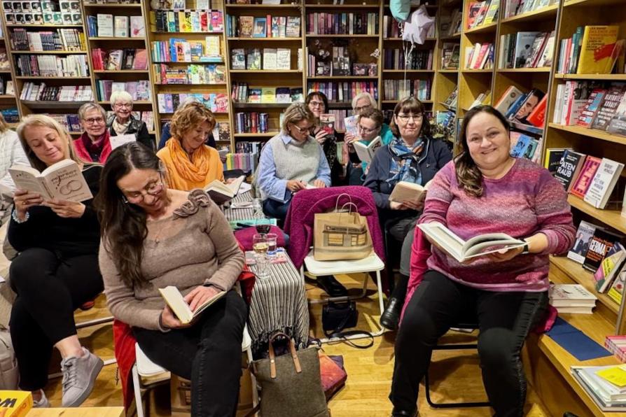 9 verschiedene Frauen sitzung auf Stühlen in der Buchhandlung. Einige haben Bücher in der Hand und lesen in diesen, andere schauen lächelnd in die Kamera. Der Hintergrund ist gefüllt mit Deckenhohen Bücherregalen, in denen Bücher verschiedener Genres zum Erwerb stehen.