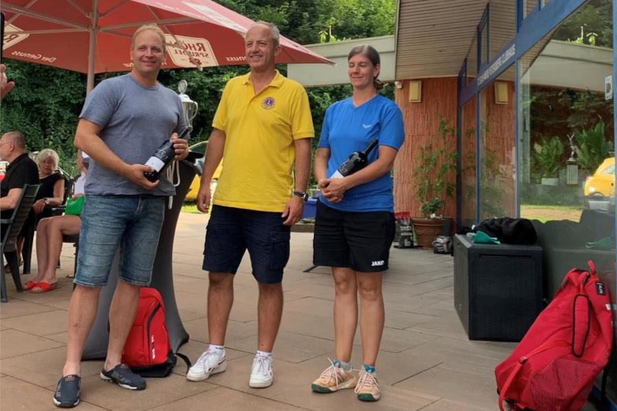 Zu sehen sind 2 Herren und eine Dame in sportlicher Kleidung. Sie befinden sich auf der Terasse eines Vereinsheims am Sportplatz. Der Herr links und die Dame rechts sind die Siegenden des Turniers und halten jeweils eine Flasche Wein in der Hand. Der Herr in der Mitte ist der aktuelle Clubpräsident, der in die Kamera lächelt. Gut zu erkennen, an seinem gelben Shirt mit Lions Logo.